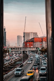 Heavy traffic flows along a multi-lane highway in an urban setting, bordered by tall buildings under construction, with several cranes visible against a sunset sky.
