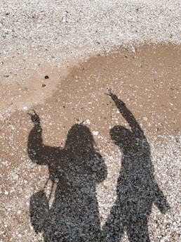Two shadows are cast on a textured ground, with one shadow showing a figure pointing upwards and the other also appearing to gesture. The surface is a mix of small pebbles and sandy patches.