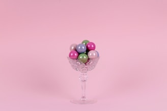 A crystal glass filled with colorful, glossy spherical candies is set against a soft pink background. The glass has an intricate cut pattern and a long stem.