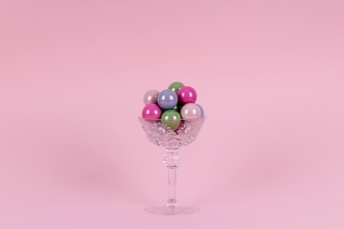 A crystal glass filled with colorful, glossy spherical candies is set against a soft pink background. The glass has an intricate cut pattern and a long stem.