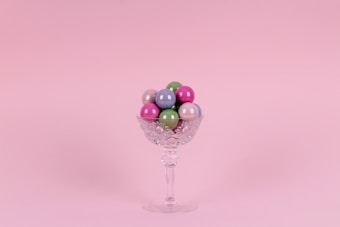 A crystal glass filled with colorful, glossy spherical candies is set against a soft pink background. The glass has an intricate cut pattern and a long stem.
