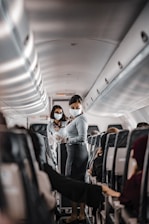 Two flight attendants wearing masks are in the aisle of an airplane. They are interacting with passengers while maintaining safety protocols. The seating area is visible with rows of occupied seats, and the interior lighting is subtle.