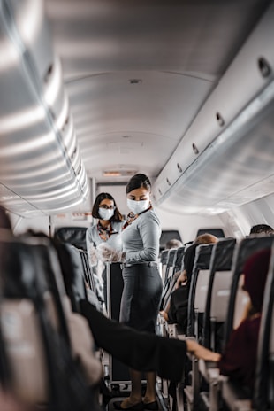 Two flight attendants wearing masks are in the aisle of an airplane. They are interacting with passengers while maintaining safety protocols. The seating area is visible with rows of occupied seats, and the interior lighting is subtle.
