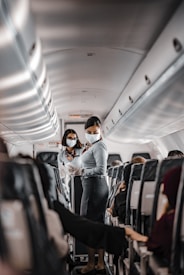 Two flight attendants wearing masks are in the aisle of an airplane. They are interacting with passengers while maintaining safety protocols. The seating area is visible with rows of occupied seats, and the interior lighting is subtle.