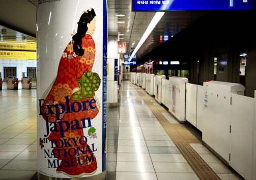 A subway station scene featuring a large pillar poster advertising the Tokyo National Museum with the text 'Explore Japan'. The poster depicts a traditional Japanese figure in a vibrant red kimono adorned with floral patterns, standing out against the modern, clean station interior. The station's tiled floor and signage in multiple languages are visible, along with the tracks and platform barriers.