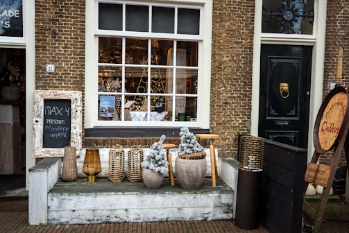 A charming storefront with large windows showcasing decorative items inside a shop. The exterior features brick walls and a dark green door with a decorative knocker. In front of the window, various objects such as a chalkboard sign, potted plants, lanterns, and wicker baskets are arranged. Nearby, there is a large round wooden sign and more decorative items, adding a rustic and inviting feel.