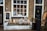A charming storefront with large windows showcasing decorative items inside a shop. The exterior features brick walls and a dark green door with a decorative knocker. In front of the window, various objects such as a chalkboard sign, potted plants, lanterns, and wicker baskets are arranged. Nearby, there is a large round wooden sign and more decorative items, adding a rustic and inviting feel.