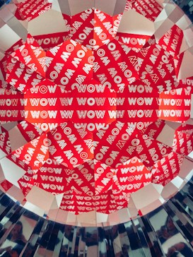 A colorful and geometric pattern featuring the word 'WOW' repeated multiple times in white on a red background. The arrangement appears kaleidoscopic, creating a vibrant and eye-catching visual effect.