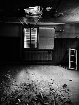 An abandoned room with a damaged ceiling showing exposed beams and debris scattered across the wooden floor. A window, partially covered with boards, allows some light to enter the otherwise dark space. A leaning ladder rests against the wall next to a radiator beneath the window.
