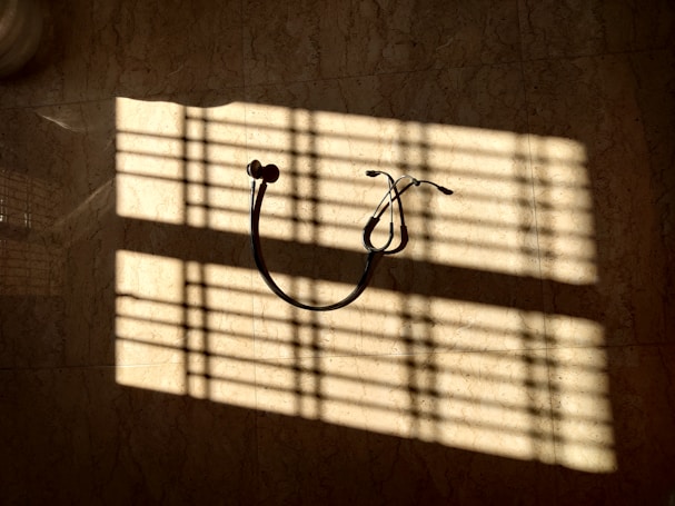 A stethoscope hangs on a wall, illuminated by sunlight streaming through blinds, casting striped shadows across the surface.