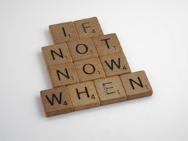 Wooden Scrabble tiles spell out the motivational phrase 'If not now, when' arranged in a pyramid shape on a light background.