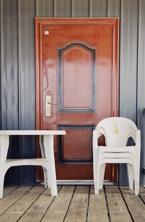 A wooden door with a deep brown color is framed by vertical gray panels. In front of the door, there is a white plastic table and three stacked white plastic chairs with a yellow logo or design on the top chair. The flooring is made of wooden planks, adding a rustic touch to the scene.