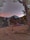 Tents are set up in a rustic campsite surrounded by tall trees. A signpost with arrows is visible, pointing in different directions. The background features a mountain silhouette against a colorful sky, suggesting either sunrise or sunset. Several people are scattered around the campsite area.