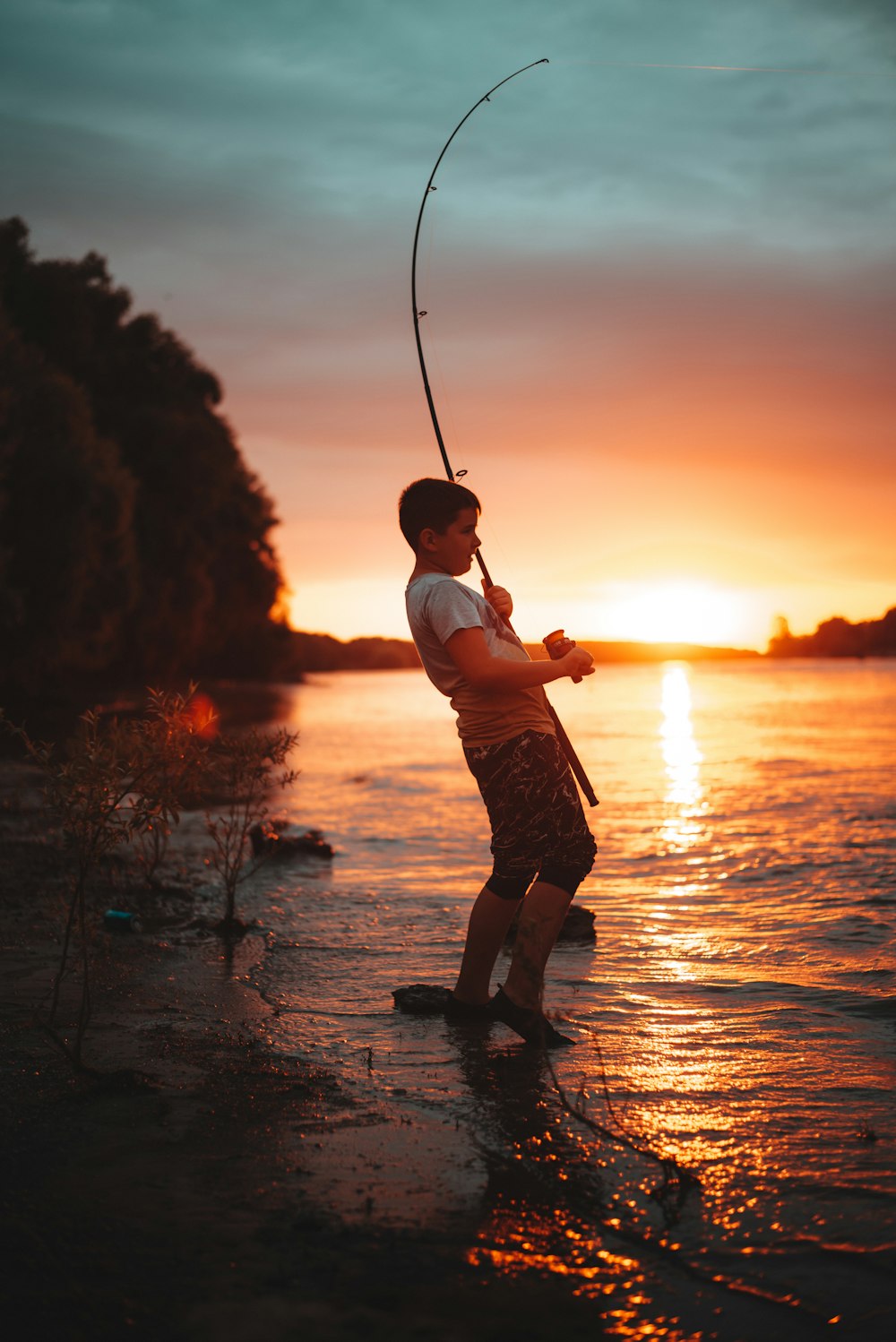 menino na camisa branca e shorts marrons segurando vara de pesca durante o pôr do sol