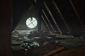 A dimly lit attic space with a large circular window that has a cross-shaped design. The floor is cluttered with scattered books and debris, creating a sense of abandonment and neglect. Sunlight filters through the dusty window, casting soft light across the room.
