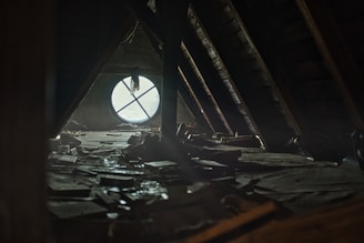 A dimly lit attic space with a large circular window that has a cross-shaped design. The floor is cluttered with scattered books and debris, creating a sense of abandonment and neglect. Sunlight filters through the dusty window, casting soft light across the room.