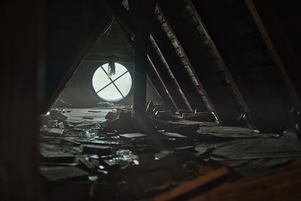 A dimly lit attic space with a large circular window that has a cross-shaped design. The floor is cluttered with scattered books and debris, creating a sense of abandonment and neglect. Sunlight filters through the dusty window, casting soft light across the room.