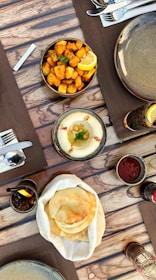 An overhead view of a rustic dining table setup with various dishes and drinks. There is a bowl of seasoned roasted potatoes garnished with a lemon wedge and parsley. Nearby, there is a plate of hummus topped with olive oil and spices. A basket of flatbreads is placed in the center. Drinks served with lemon wedges and a bottle of soda are also present.