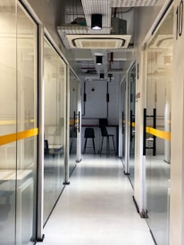 A modern office hallway with glass partition walls and a clean, minimalist design. The corridor is lit with overhead lights, leading to a small seating area with two high stools at a bar-style table. The walls are aligned with visible ceiling fixtures and white and yellow color accents.