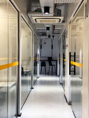 A modern office hallway with glass partition walls and a clean, minimalist design. The corridor is lit with overhead lights, leading to a small seating area with two high stools at a bar-style table. The walls are aligned with visible ceiling fixtures and white and yellow color accents.