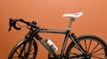 A sleek racing bicycle with a black frame and white seat is set against a plain brown background. It features drop handlebars and thin tires, suitable for speed and aerodynamic efficiency. A water bottle is mounted on the frame.