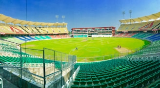 A large, empty cricket stadium with tiered seating in various colors surrounds a well-maintained green field. Tall floodlights are positioned at the top of the stadium, and some maintenance personnel are working on the field. The sky is clear and bright.