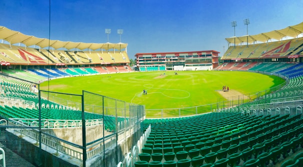A large, empty cricket stadium with tiered seating in various colors surrounds a well-maintained green field. Tall floodlights are positioned at the top of the stadium, and some maintenance personnel are working on the field. The sky is clear and bright.