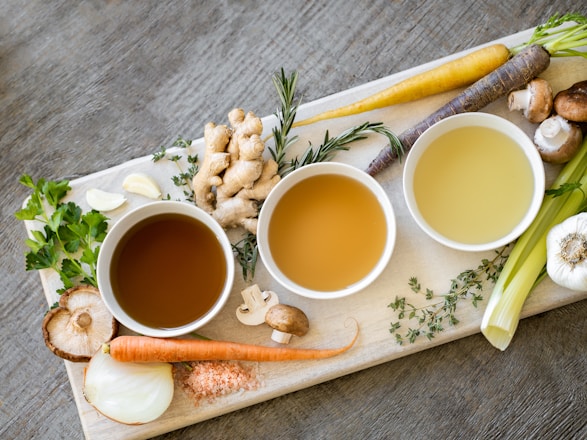 A rustic wooden table with steaming bowls of bone broth surrounded by fresh herbs and natural ingredients.