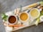 Three bowls containing different broths are placed on a wooden chopping board alongside a variety of fresh vegetables and herbs, including carrots, celery, garlic, mushrooms, ginger, and rosemary. There is also salt and onions present.