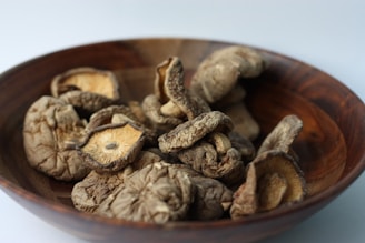 A wooden bowl contains several dried mushrooms. The mushrooms have a shriveled and wrinkled texture, with a mix of light brown and dark brown colors. The bowl itself is made of wood, featuring visible grain patterns.