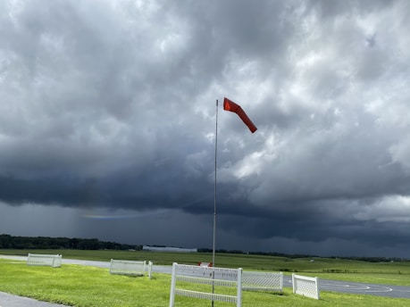 A windsock stands upright in the middle of a green field, indicating windy conditions. Dense, dark clouds fill the sky, suggesting an approaching storm. The landscape is flat, with patches of sunlight breaking through to illuminate parts of the grass.