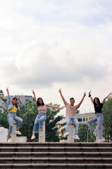 Four people are energetically posing on top of concrete posts with their arms raised and making peace signs. They are smiling and appear to be enjoying themselves, indicating a vibrant and joyful moment. The background includes trees and a building with colorful windows.