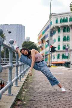A person is doing a stretching exercise, with one leg raised and placed on a railing along a sidewalk. The background features a multi-colored building and urban cityscape. The individual is casually dressed in a sleeveless top and jeans, with white sneakers.