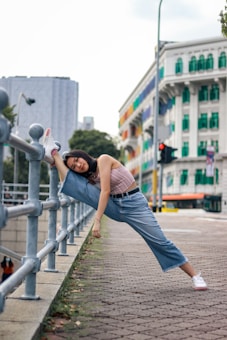 A person is doing a stretching exercise, with one leg raised and placed on a railing along a sidewalk. The background features a multi-colored building and urban cityscape. The individual is casually dressed in a sleeveless top and jeans, with white sneakers.