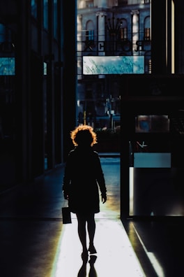 A silhouette of a person with curly hair walking through a dimly lit hallway. The backlighting creates a striking contrast, highlighting their hair and casting a long shadow on the floor. In the background, a building facade and signs are faintly visible.