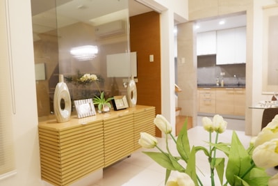 A modern and elegantly decorated living space with a sleek wooden sideboard featuring decorative items such as vases and picture frames. In the foreground are fresh white tulips, adding a touch of nature to the interior. The background shows a kitchen area with wooden cabinets and a gray countertop, creating a warm and inviting atmosphere.