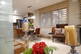 A modern and cozy living space featuring a dining table with four chairs, partially visible behind elegant sheer blinds that allow natural light to filter through. The room is decorated with lush plants and candles, enhancing its warm and inviting atmosphere. A comfortable seating area is adorned with decorative pillows, and a well-organized kitchen area can be seen in the background.