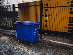 blue trash bin beside yellow metal fence