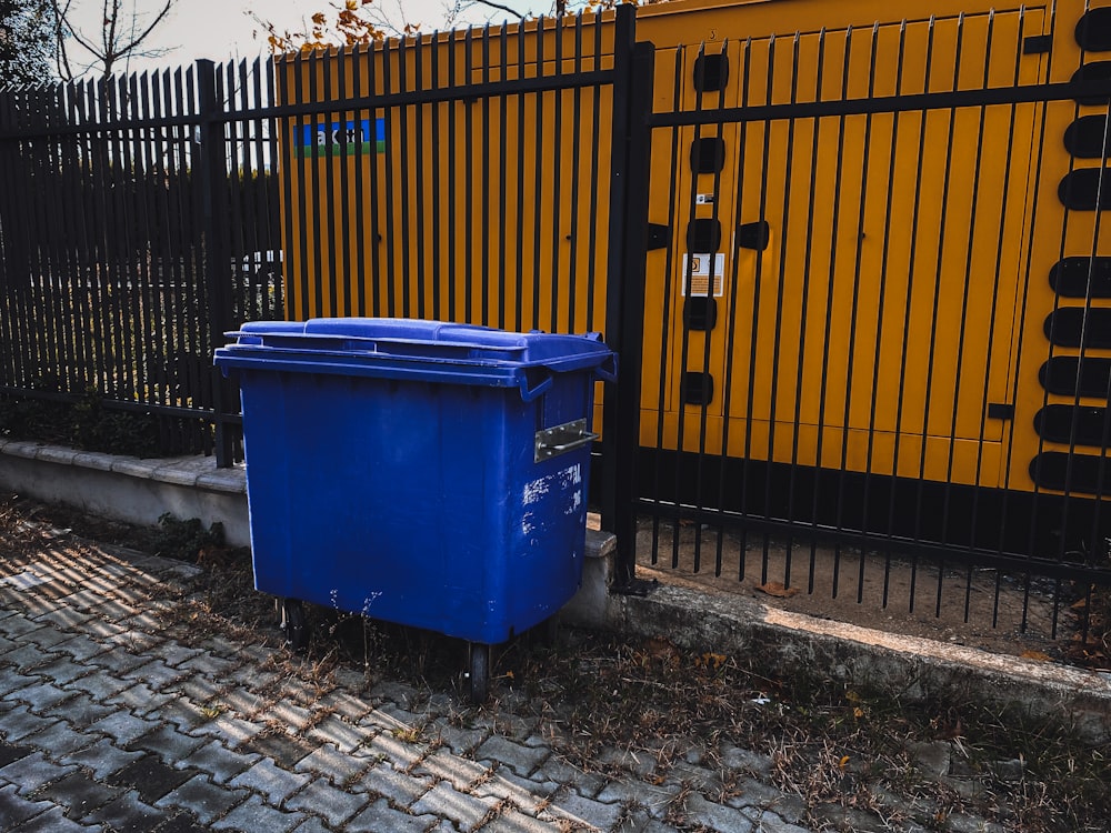 blue trash bin beside yellow metal fence