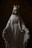 A statue of a woman wearing a flowing robe is adorned with a golden crown. The figure's arms are extended slightly with palms facing outward. A chain or rosary is draped over the shoulders, with a dark, textured background providing contrast.