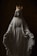 A statue of a woman wearing a flowing robe is adorned with a golden crown. The figure's arms are extended slightly with palms facing outward. A chain or rosary is draped over the shoulders, with a dark, textured background providing contrast.
