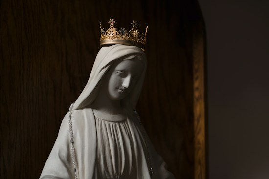 A serene statue of a woman with a gentle expression is adorned with a delicate crown on her head. The statue wears a flowing garment and a beaded rosary drapes over the figure's shoulder. Warm lighting casts soft shadows, enhancing the peaceful and sacred atmosphere.