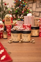 A festive scene with Christmas decorations, including a decorated tree with colorful lights. Various wrapped gifts, predominantly red and white, are arranged around products labeled as hemp extract dog chews and tinctures. The setting is cozy with a wooden table surface.