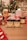 A festive scene with Christmas decorations, including a decorated tree with colorful lights. Various wrapped gifts, predominantly red and white, are arranged around products labeled as hemp extract dog chews and tinctures. The setting is cozy with a wooden table surface.