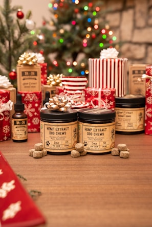 A festive scene with Christmas decorations, including a decorated tree with colorful lights. Various wrapped gifts, predominantly red and white, are arranged around products labeled as hemp extract dog chews and tinctures. The setting is cozy with a wooden table surface.