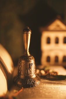 A vintage silver bell with ornate designs is placed on an open book. In the background, there is a blurred miniature house or building model, lending a warm, nostalgic atmosphere.