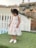 A young child is wearing a white lace dress with floral patterns and white shoes, standing on a patch of artificial green grass. A white fence and a decorative stone wall are visible in the background.