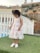 A young child is wearing a white lace dress with floral patterns and white shoes, standing on a patch of artificial green grass. A white fence and a decorative stone wall are visible in the background.