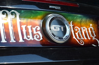 The image displays the back of a car featuring a decorative plate with the word 'Mustang' written in a stylized font. The plate has a vibrant gradient background with shades of green, orange, and red. In the center, there is a prominent circular GT emblem.