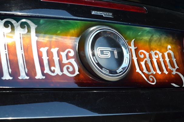 The image displays the back of a car featuring a decorative plate with the word 'Mustang' written in a stylized font. The plate has a vibrant gradient background with shades of green, orange, and red. In the center, there is a prominent circular GT emblem.